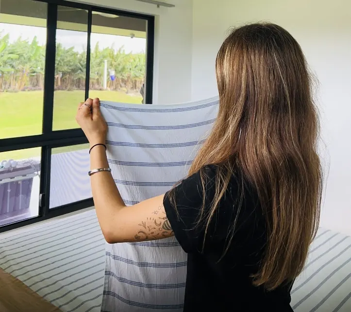Citrus cleaner shaking out fresh striped bed linen in a bright bedroom overlooking a green garden
